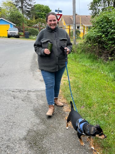 A reviewer walks their small dog on a path, with the dog wearing a blue Pawfalks™ harness.